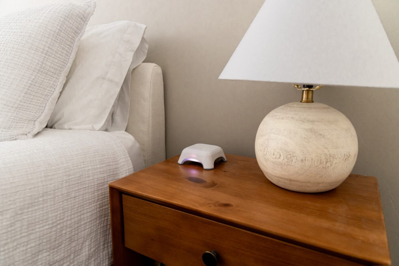 Small white BioFi sleep sensor emitting a soft glow on a wooden nightstand next to a bed and lamp