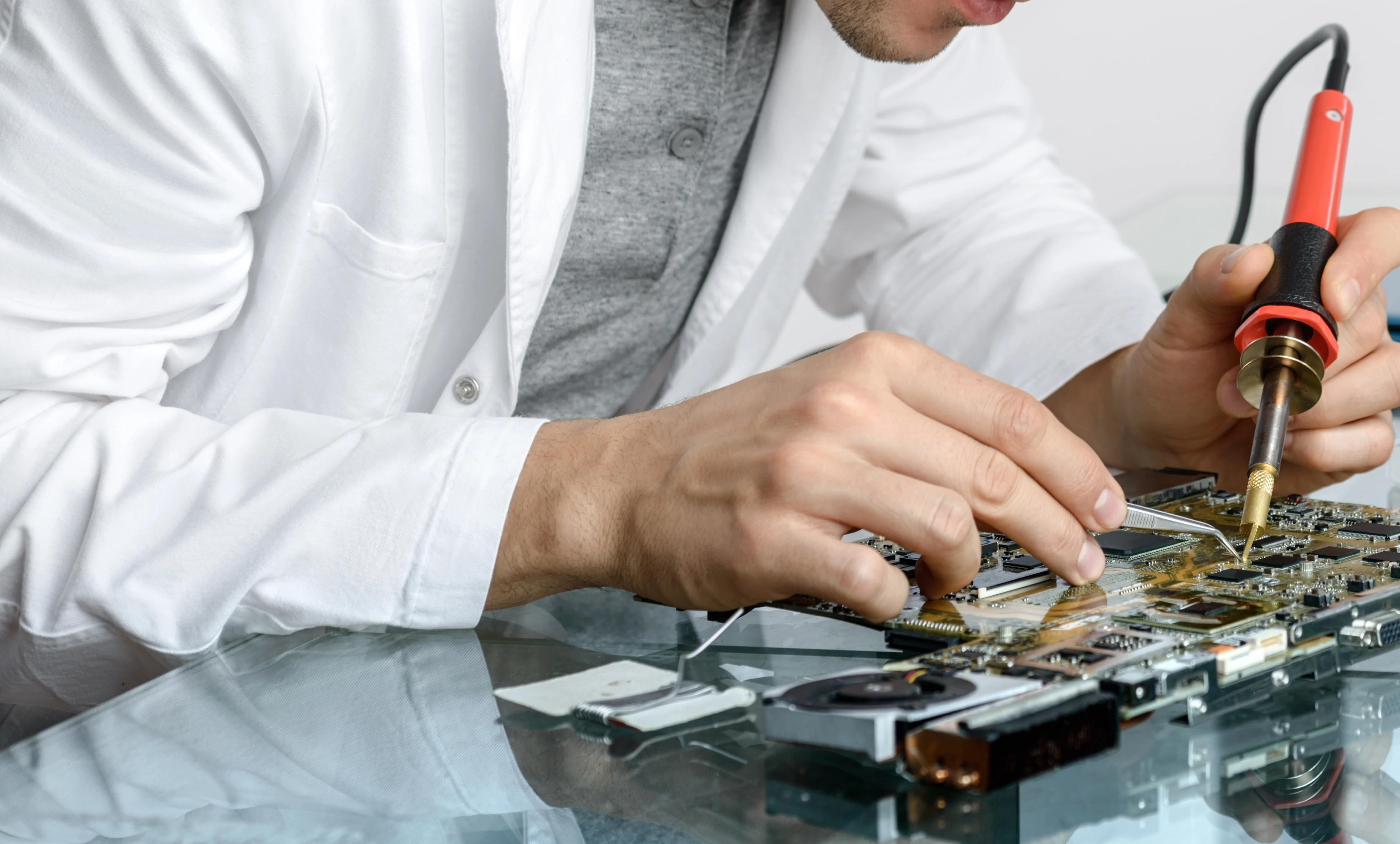 Close-up of hands using a soldering iron and tweezers to attach components onto a circuit board resting on a glass surface